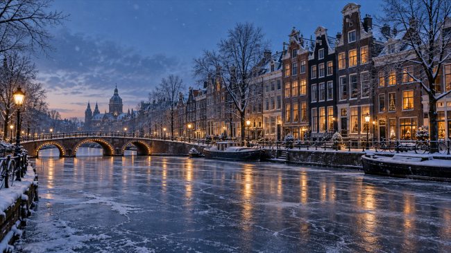 Zugefrorener Kanal mit verschneiten Giebelhäusern und beleuchteten Brücken